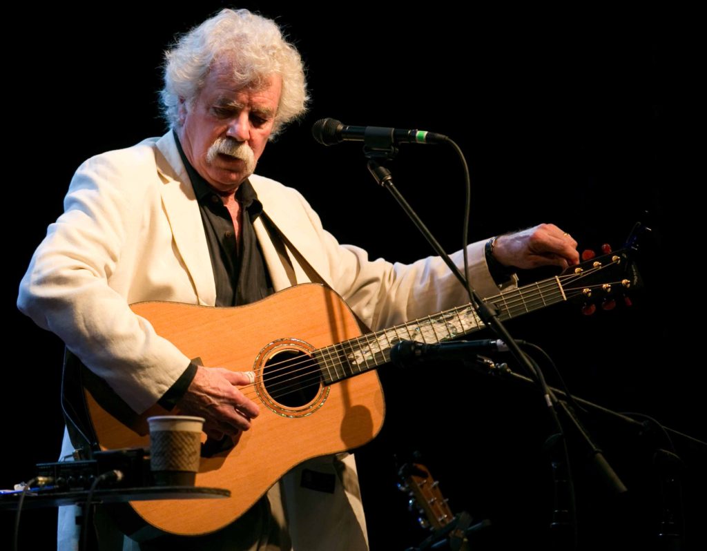 Man with white hair and mustache, Tom Rush, playing a guitar at a microphone