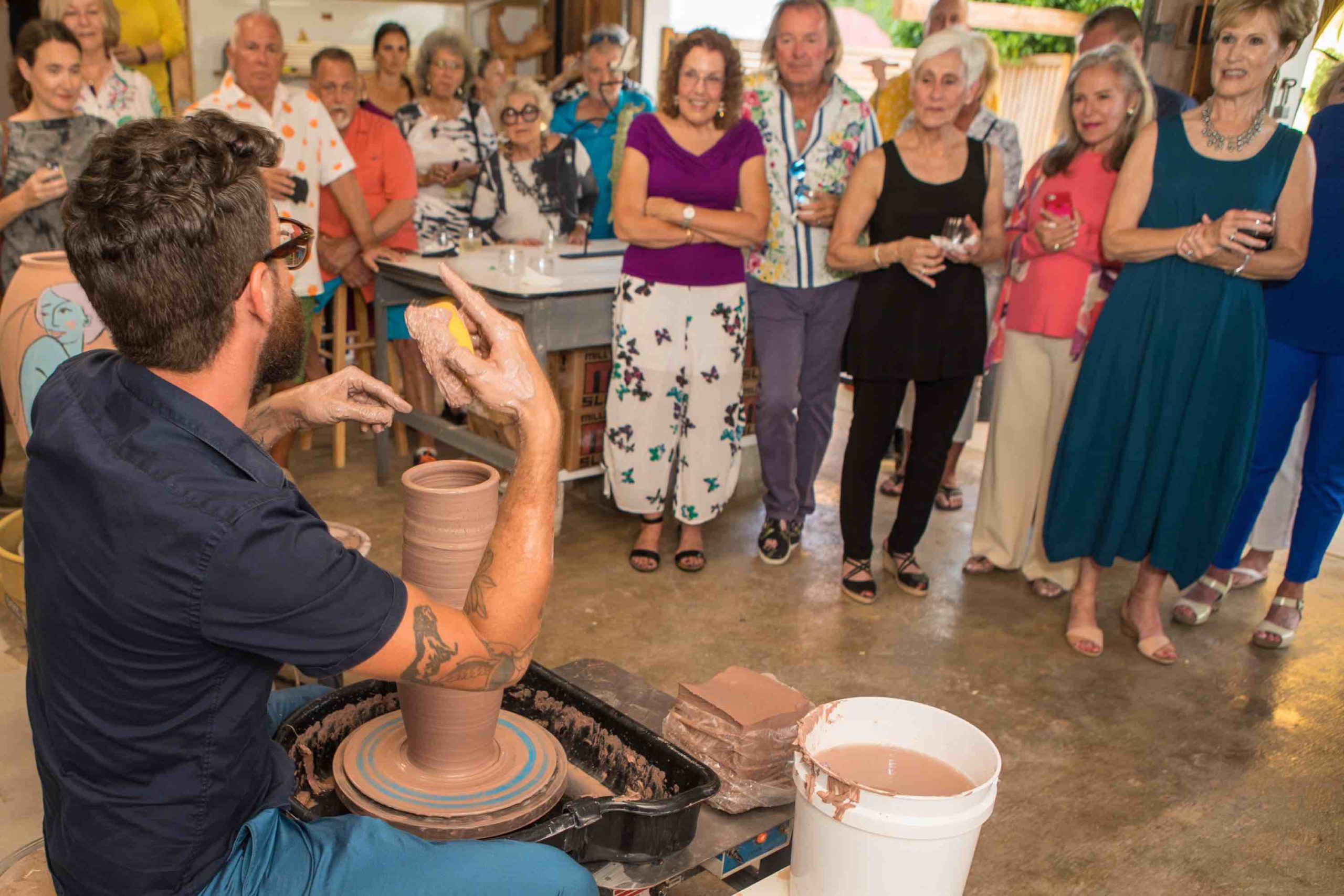 People stading watching a ceramic potter sitting at a wheel making a pot