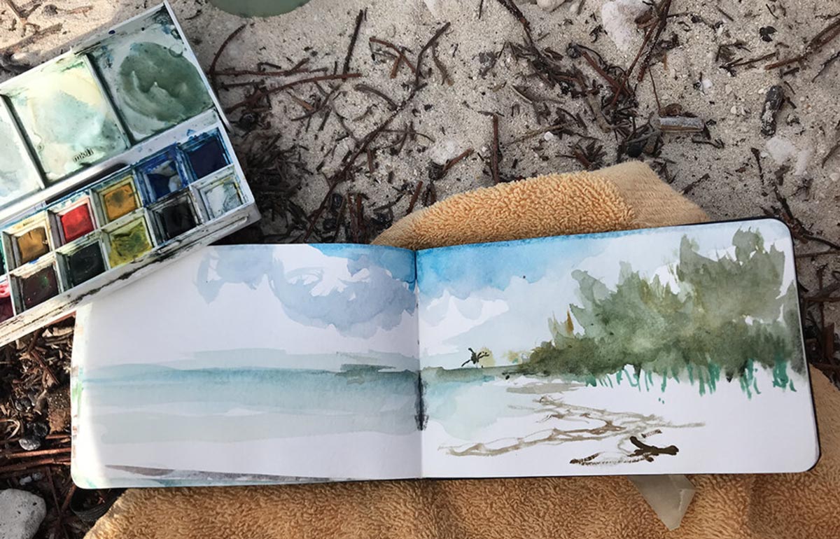 photo of view of watercolor in sketchbook on sand and watercolor paintbox
