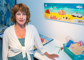 A woman in a white cardigan leaning on a table in an artist studio with paintings on the wall behind her of beach scenes