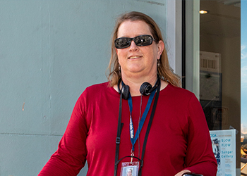 Photo of a female reporter in a bright red shirt and sunglasses with a press lanyard and recording equipment around her neck