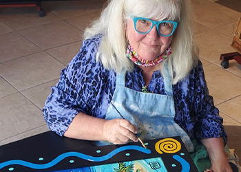 Photo of a female artist with white shoulder length hair wearing aqua glasses and painting a design on a black frame