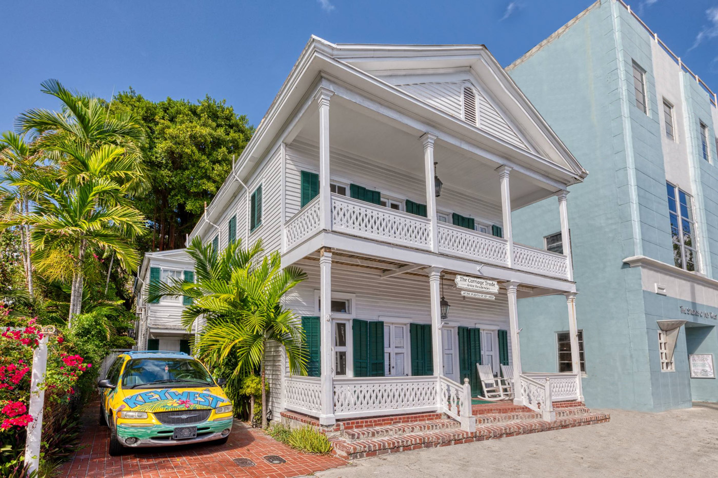 Two story white wooden house with green shutters, porches and a colorful painted car on left.