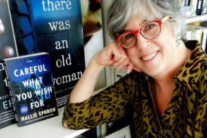 Woman In red glasses leaning on a book shelf in front of her book covers -Hallie Ephron