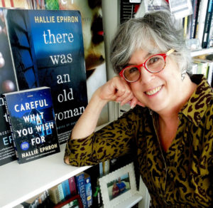 Woman In red glasses leaning on a book shelf in front of her book covers -Hallie Ephron
