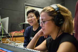 Asian woman with short black hair and eye glasses smiling with a young woman student in foreground out of focus wearing over the ear headphones and working a soundboard.