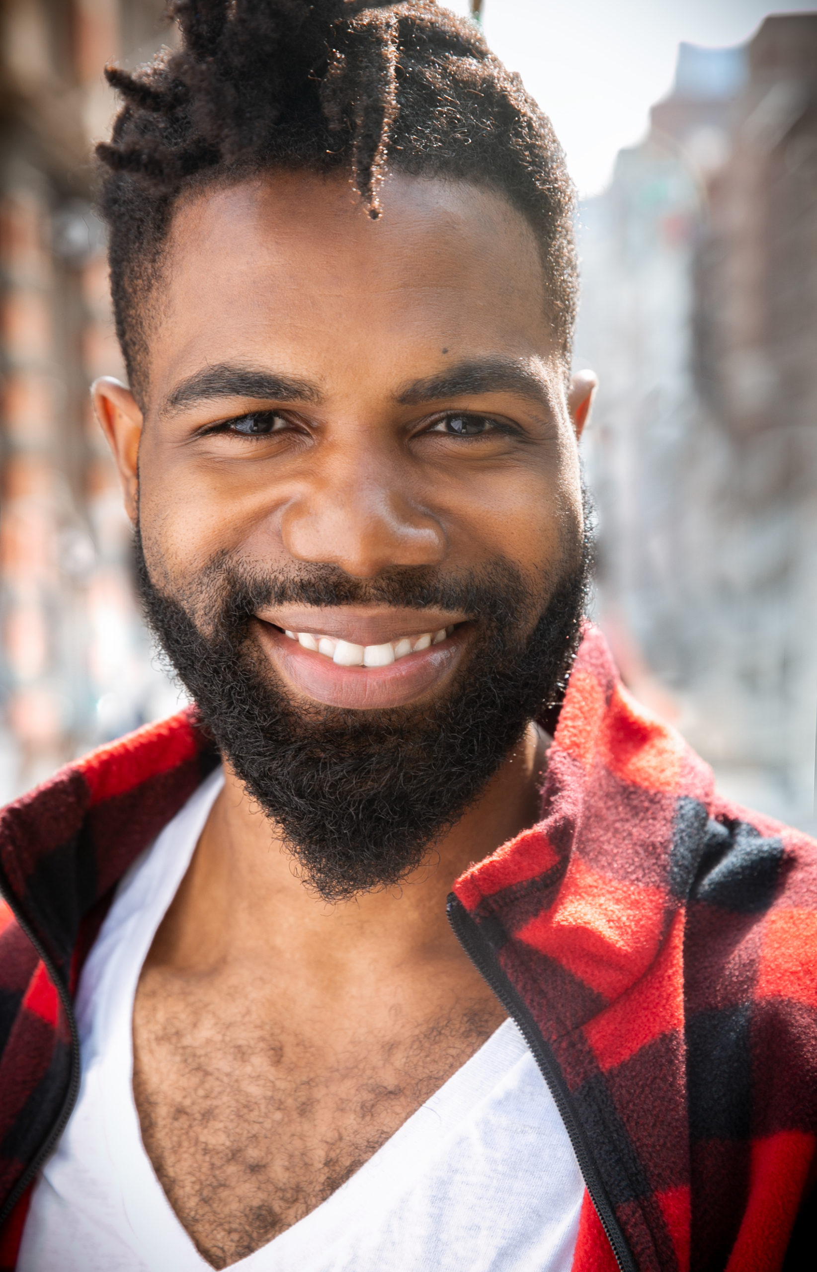 Black man in white v-neck t-shirt and red and black checkered flannel with a beard and short dreadlocked hair smiling at the camera
