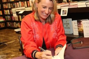 Photo of a blonde woman signing a book in a red jacket