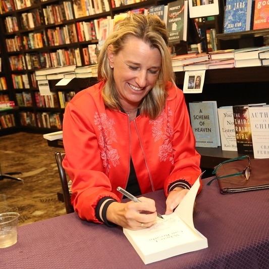 Photo of a blonde woman signing a book in a red jacket