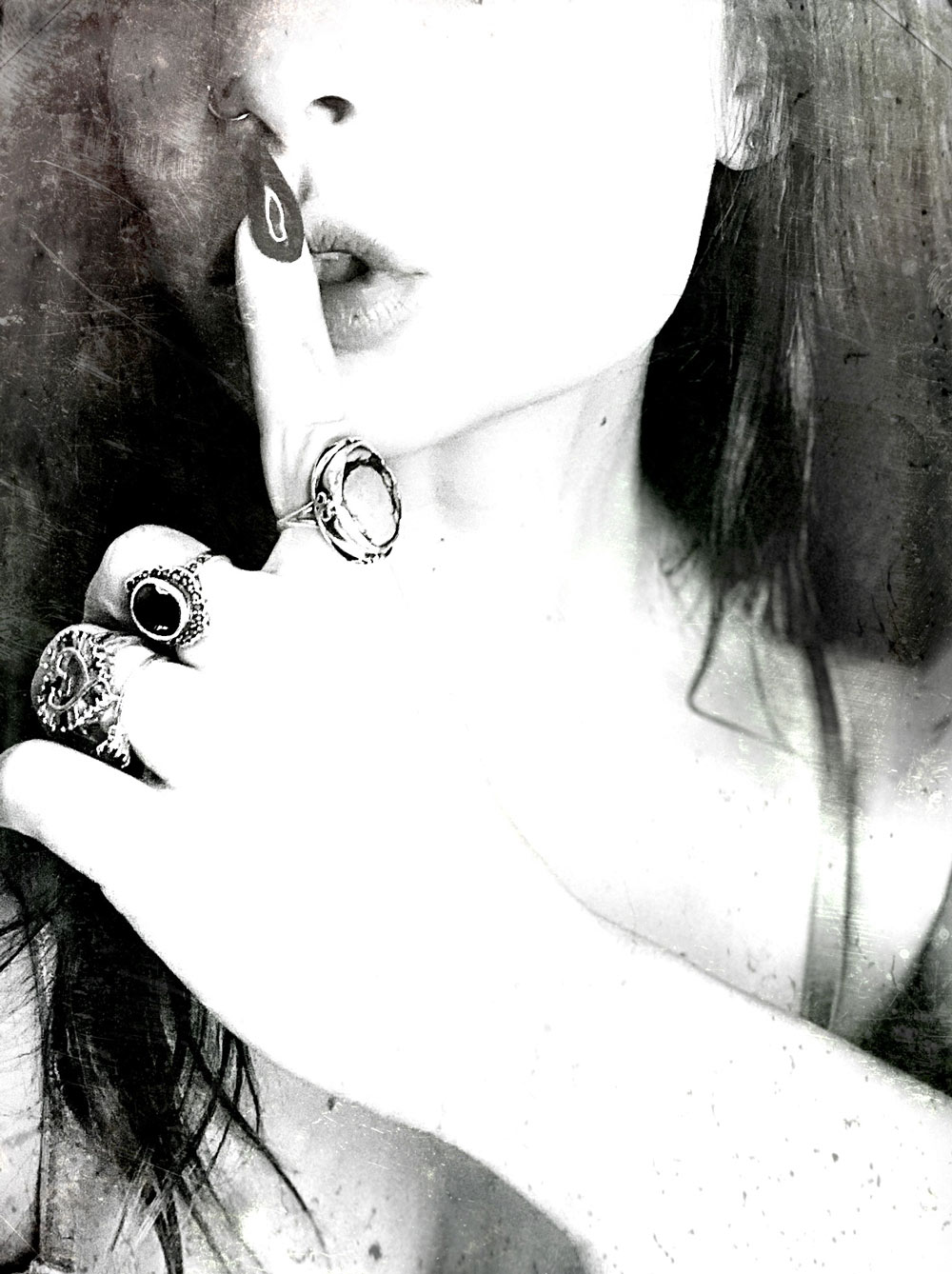 Black and white photo of a woman's hand wearing chunky rings shushing someone (holding her pointer finger in front of her lips).