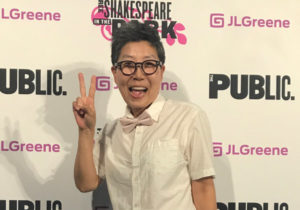 Asian woman with short dark brown hair and glasses smiling and holding a peace sign in front of a red carpet backdrop for SHAKESPEARE IN THE PARK.