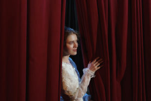 Photo of a brunette woman in a tulle long sleeved top peeking out from behind a red curtain on stage.