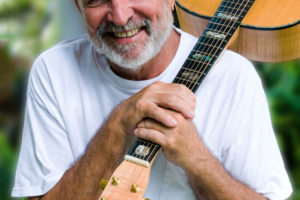 Man smiling with grey beard in a white t-shirt and jeans holding a guitar over his shoulder.