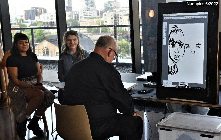 Photo of an artist drawing caricatures of two young women on a surface being transcribed onto a computer screen.