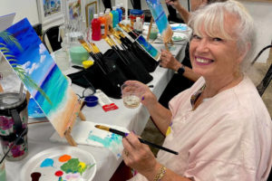 Woman smiling whole holding a paintbrush and glass of wine painting a tropical beach scene.