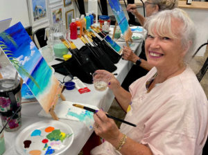 Woman smiling whole holding a paintbrush and glass of wine painting a tropical beach scene.