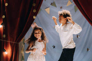 A young girl and boy jumping and smiling on stage with gold stars and discs behind and a red stage curtain.