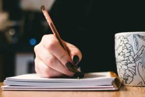 Woman's hand writing with a pen in a notebook with long black nails and a coffee mug on the right.