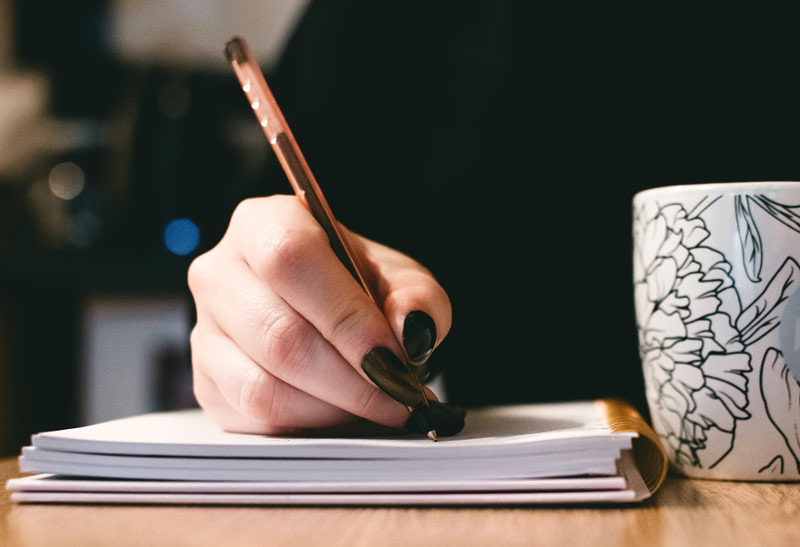 Woman's hand writing with a pen in a notebook with long black nails and a coffee mug on the right.