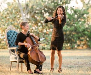 A woman seated in a blue chair playing a cello next to a woman standing playing a violin.
