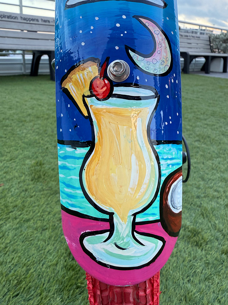 A hand-painted image of a piña colada in a curved glass with a pineapple wedge and cherry on top, set against a blue night sky with a crescent moon and an ocean horizon.