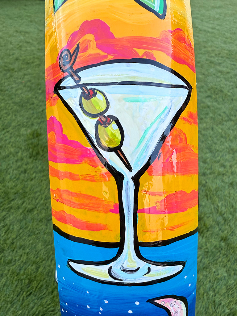 A hand-painted martini with two olives on a skewer, shown against a bright orange and pink sunset sky above a strip of blue water.