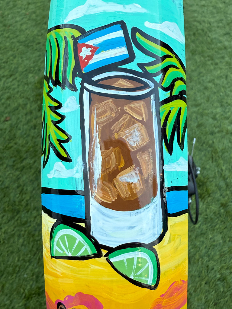 A tall iced rum cocktail with a small Cuban flag, surrounded by palm fronds and lime wedges, with a turquoise sky and ocean in the background.