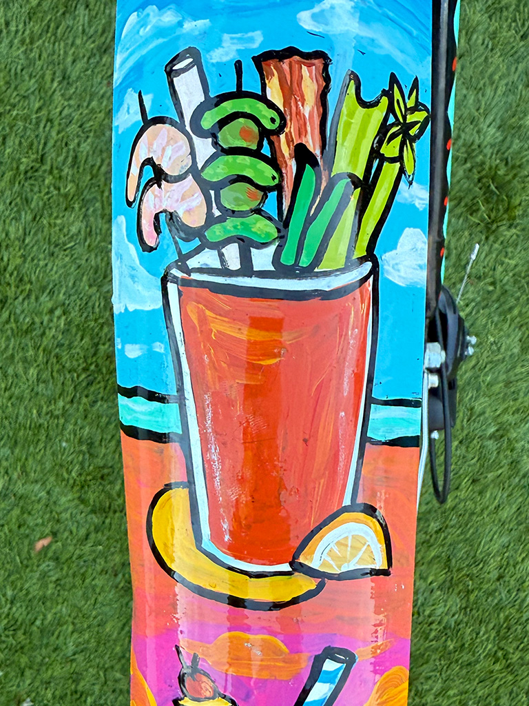 A hand-painted Bloody Mary in a tall red glass overflowing with garnishes—shrimp, olives, celery, and bacon—set against a bright beachy blue sky and ocean.