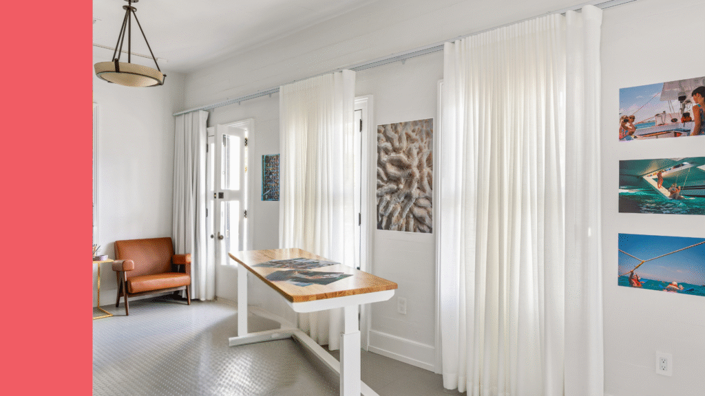 Bright interior of the Peyton Evans Artist Residency in Key West with white curtains, art on the walls, a modern wood table, and a leather chair.
