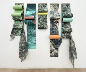 Installation artwork titled “10 años fuera de casa” by Miami-based artist Verónica Pasman. Five vertical scrolls of marbled photographic prints hang side by side on a white wall, each layered with soft fabric and pastel-colored cylinders in green, pink, yellow, turquoise, and orange. The cascading forms evoke movement and water, blending photography, fabric, and sculptural elements.