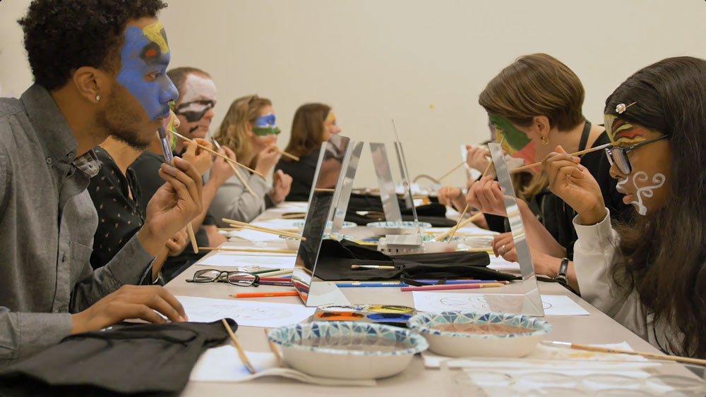 A group of people sit at a long table with mirrors, painting colorful designs on their own faces during a creative workshop.