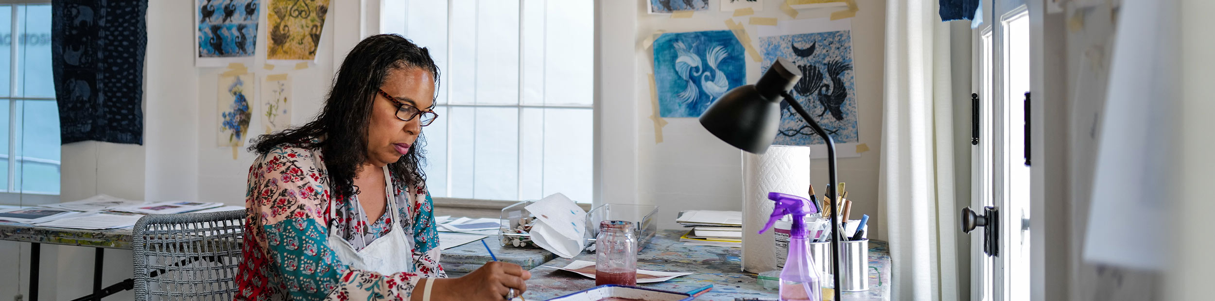 PEAR-by-michael-marrero-2 A woman wearing glasses and a floral shirt sits at a paint-covered desk, concentrating as she works with a paintbrush in a light-filled studio. Artwork is taped to the walls behind her.