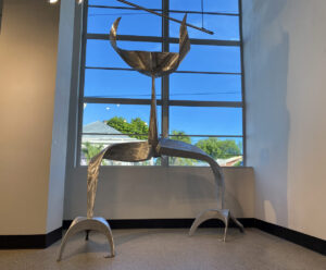 Polished metal abstract sculpture with curved, tripod-like legs and a crescent-shaped form on top, displayed in front of a tall window with natural light.