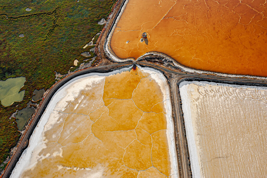 Overhead view of geometric salt ponds in orange, white, and brown tones divided by roads, with cracked textures across the surfaces.