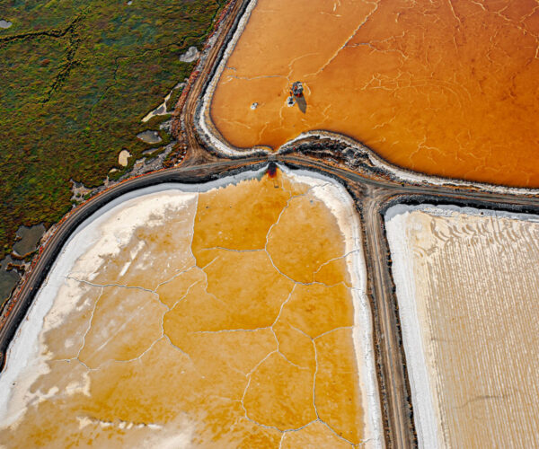 Overhead view of geometric salt ponds in orange, white, and brown tones divided by roads, with cracked textures across the surfaces.