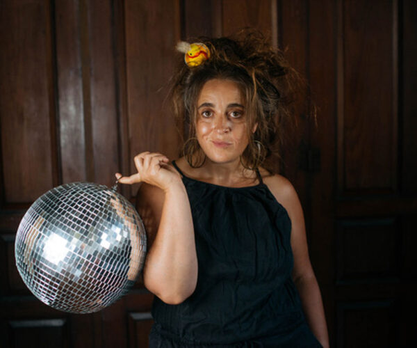 donna A woman in a black dress with messy, teased hair decorated with a small toy duck gazes at the camera while holding a shiny disco ball on one finger, standing against a dark wooden paneled background.