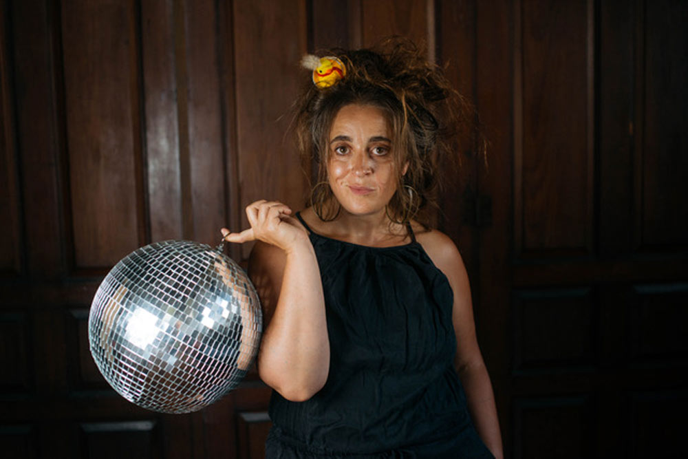 A woman in a black dress with messy, teased hair decorated with a small toy duck gazes at the camera while holding a shiny disco ball on one finger, standing against a dark wooden paneled background.