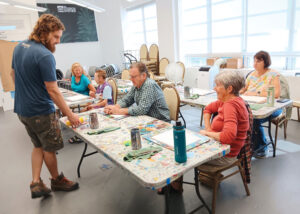 An art instructor stands at the head of a table, speaking with a group of adult students seated around worktables covered in paint splatters and supplies during a painting class at The Studios of Key West.