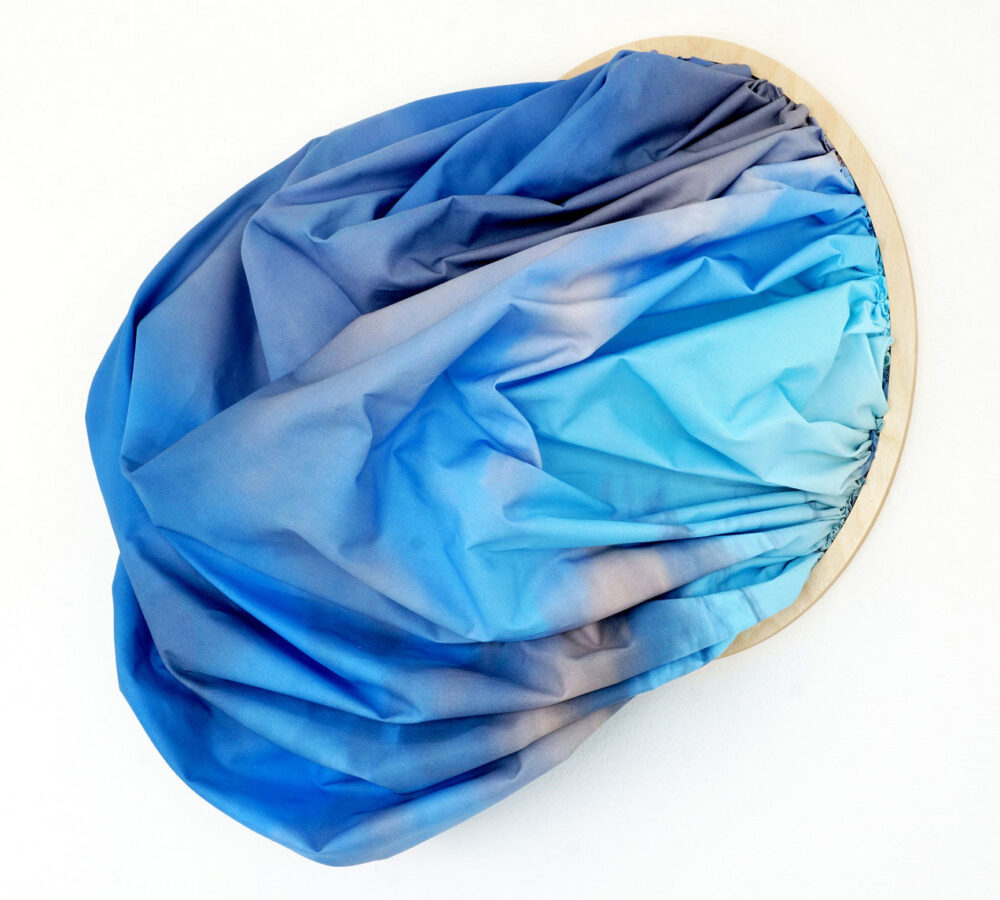 Contemporary textile artwork of gathered blue and purple fabric stretched over a circular frame, resembling ocean waves.