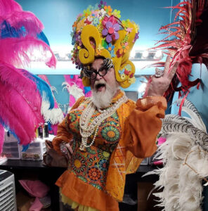 Portrait of a flamboyant performer in a colorful costume, pearl necklaces, and an oversized decorative headpiece, surrounded by feathers.