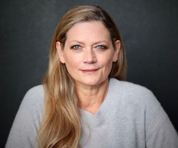 309A8446 Portrait of author Sophie Ward, wearing a light gray sweater, with long blonde hair over one shoulder, looking directly at the camera against a dark background.