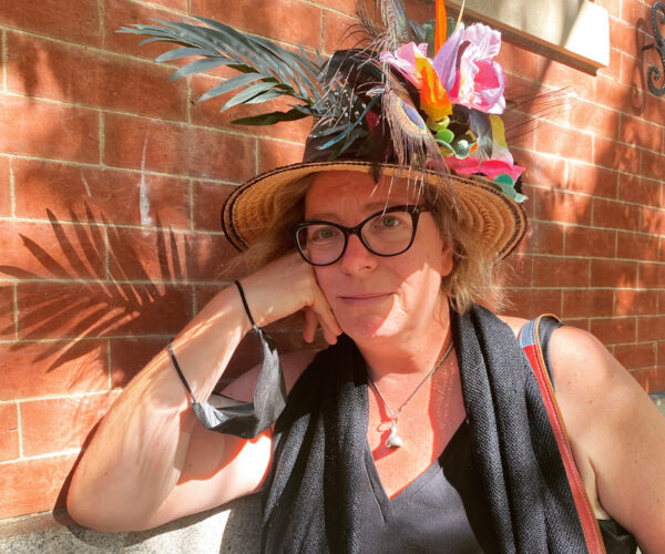 BELL_PHOTO1 Artist Kelley Bell wearing glasses and a wide-brimmed straw hat decorated with colorful flowers and feathers, leaning against a brick wall in sunlight.