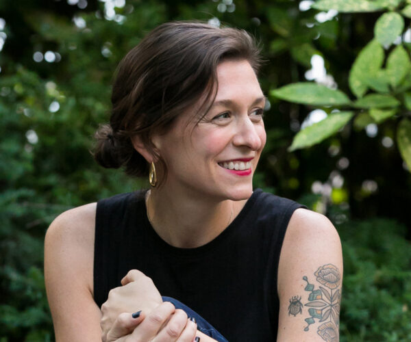 CJHauser110620_410BeowulfSheehan A person with short brown hair tied back, wearing gold hoop earrings and a black sleeveless top, smiles while seated outdoors against a backdrop of green leaves. A tattoo of flowers and a beetle is visible on their upper arm.
