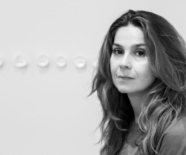 Eggs-182 A black-and-white portrait of artist Esther Sziksz, shown from the waist up. She has long, wavy hair and wears a simple top, gazing directly at the camera against a minimalist background with small, rounded forms displayed on the wall behind her.