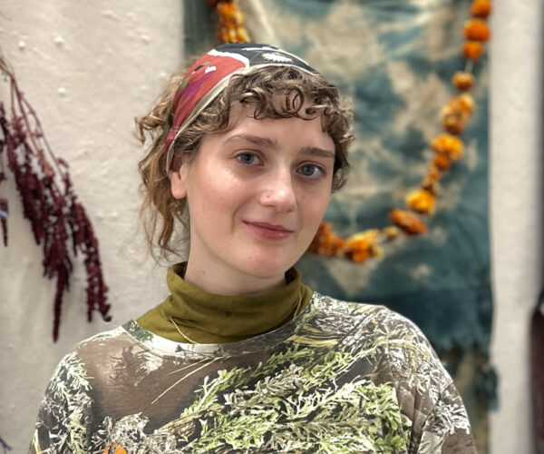 Headshot-Katherine-Miller Portrait of artist Katherine Miller, smiling slightly with a colorful headscarf and patterned shirt, standing in front of a wall with hanging textiles and marigold garland.