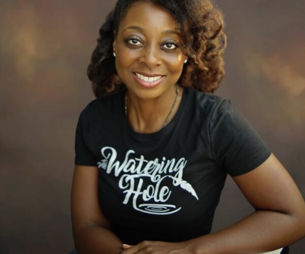 Headshot Wiley 2024.08 Credit Mingsheng He Portrait of Candace Wiley smiling, wearing a black t-shirt with 'The Watering Hole' logo, seated against a warm-toned studio backdrop. Photo by Mingsheng He.