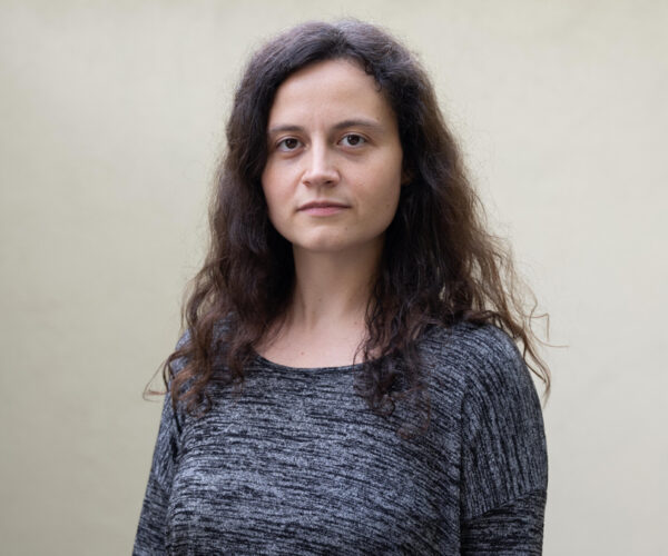 Headshot-copy A portrait of artist Sofia Valiente standing against a plain light background. She has long, wavy dark hair and wears a gray, textured long-sleeve top, gazing directly at the camera with a calm, serious expression.
