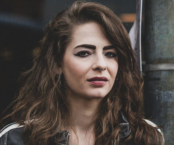 ML-RIO Author M. L. Rio with long wavy brown hair, wearing a leather jacket, photographed outdoors with a confident expression.