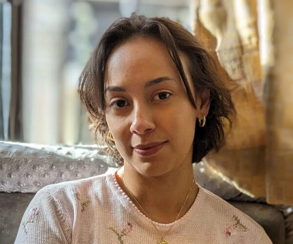 Monica-Berra-headshot-(1) Portrait of filmmaker Monica Berra, with short dark hair, smiling slightly in a neutral-toned studio setting.