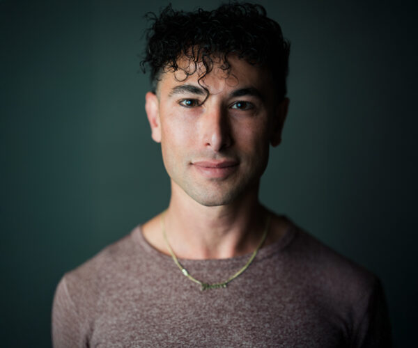 _Pioneer-Winter-photographed-by-Chantal-Lawrie-(2025) Portrait of choreographer Pioneer Winter, wearing a maroon shirt and gold necklace, looking directly at the camera with a calm expression against a dark background.